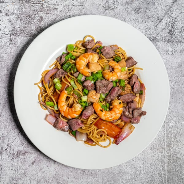 A white plate holds a vibrant dish of noodles, beef, shrimp, red onions, and green onions.
