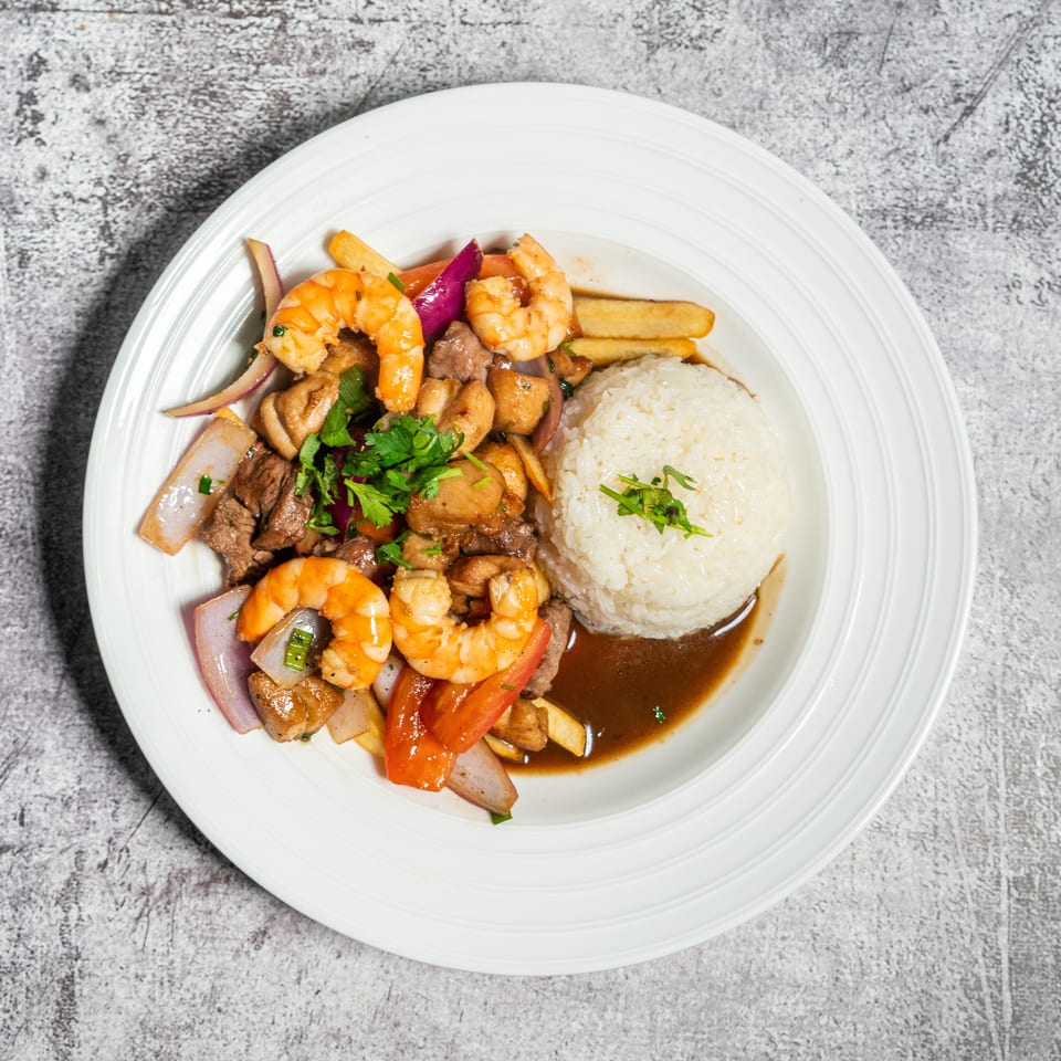 A white plate with Lomo Saltado, featuring shrimp, beef, vegetables, rice, and sauce.