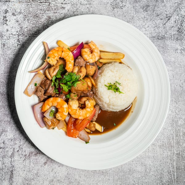 A white plate holds a dish with shrimp, beef, vegetables, rice, and a brown sauce.