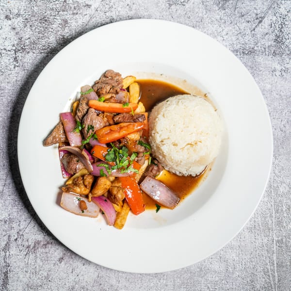 Peruvian Lomo Saltado with beef, red onions, tomatoes, fries, and rice on a white plate.